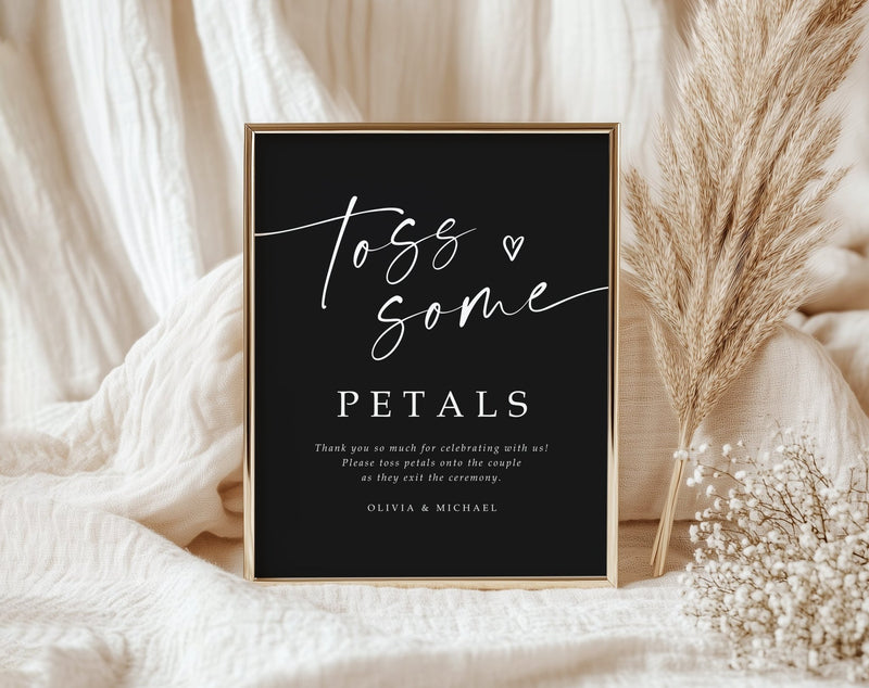 Black wedding sign with Toss Some Petals text in white calligraphy, styled with pampas grass and neutral fabric