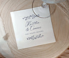 A square white paper napkin printed with the names 'Kaitlin & Conner' and 'THE NIGHT BEFORE' in dark script and floral border illustrations, placed on a round light wood slice with part of a clear wine glass visible at the top.