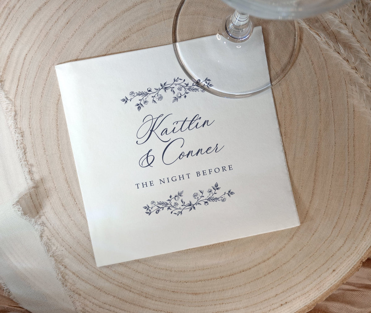 A square white paper napkin printed with the names 'Kaitlin & Conner' and 'THE NIGHT BEFORE' in dark script and floral border illustrations, placed on a round light wood slice with part of a clear wine glass visible at the top.