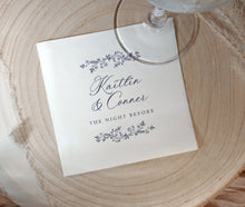 A square white paper napkin printed with the names 'Kaitlin & Conner' and 'THE NIGHT BEFORE' in dark script and floral border illustrations, placed on a round light wood slice with part of a clear wine glass visible at the top.
