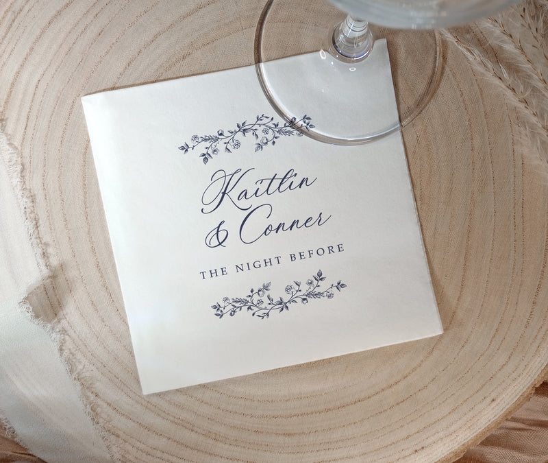 A square white paper napkin printed with the names 'Kaitlin & Conner' and 'THE NIGHT BEFORE' in dark script and floral border illustrations, placed on a round light wood slice with part of a clear wine glass visible at the top.