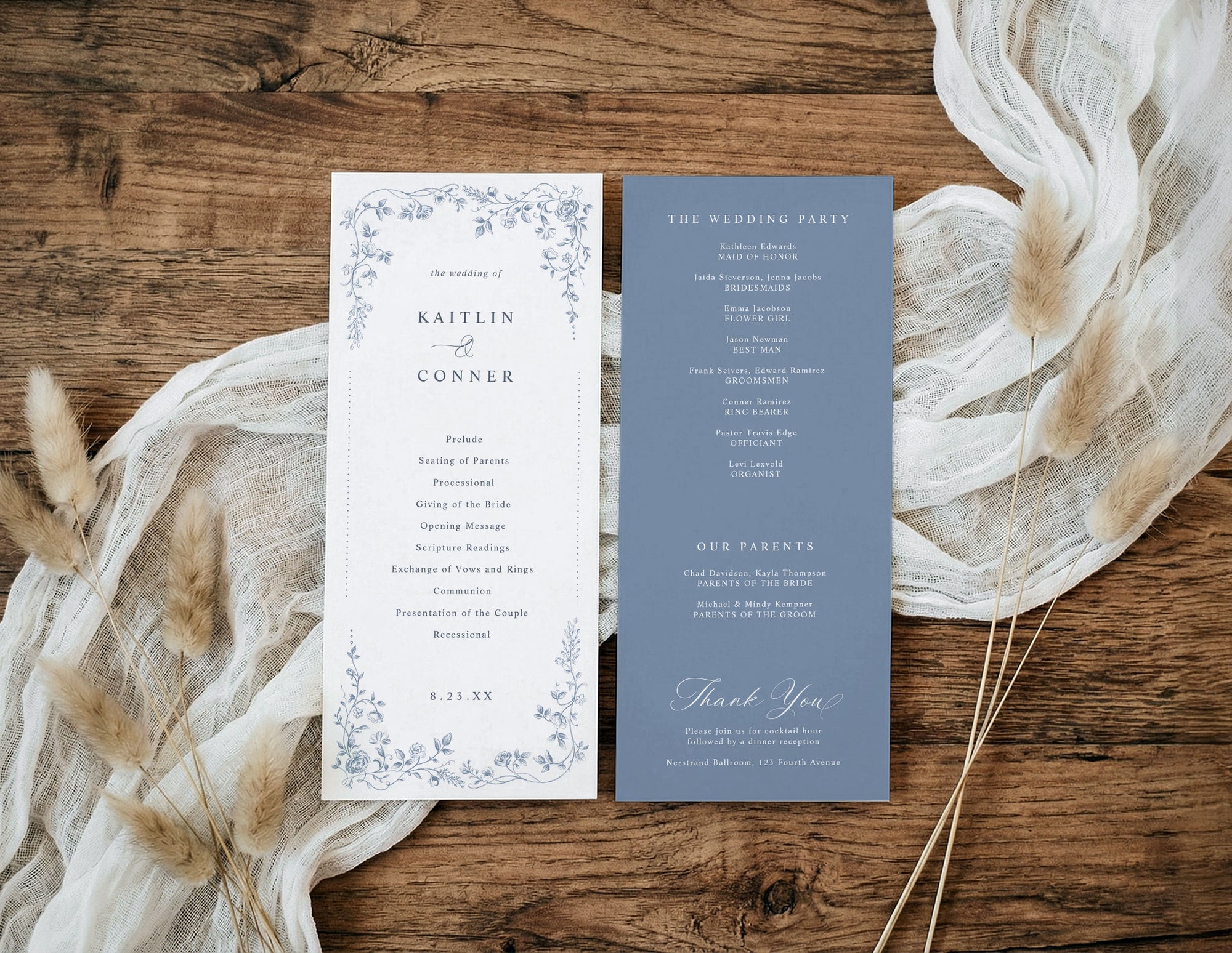 A flat-lay of a wedding program set on a rustic wooden surface, styled with cream gauze fabric and dried pampas grass. The left card is white with a delicate blue floral border and black text, while the right card is a muted slate blue with white text.