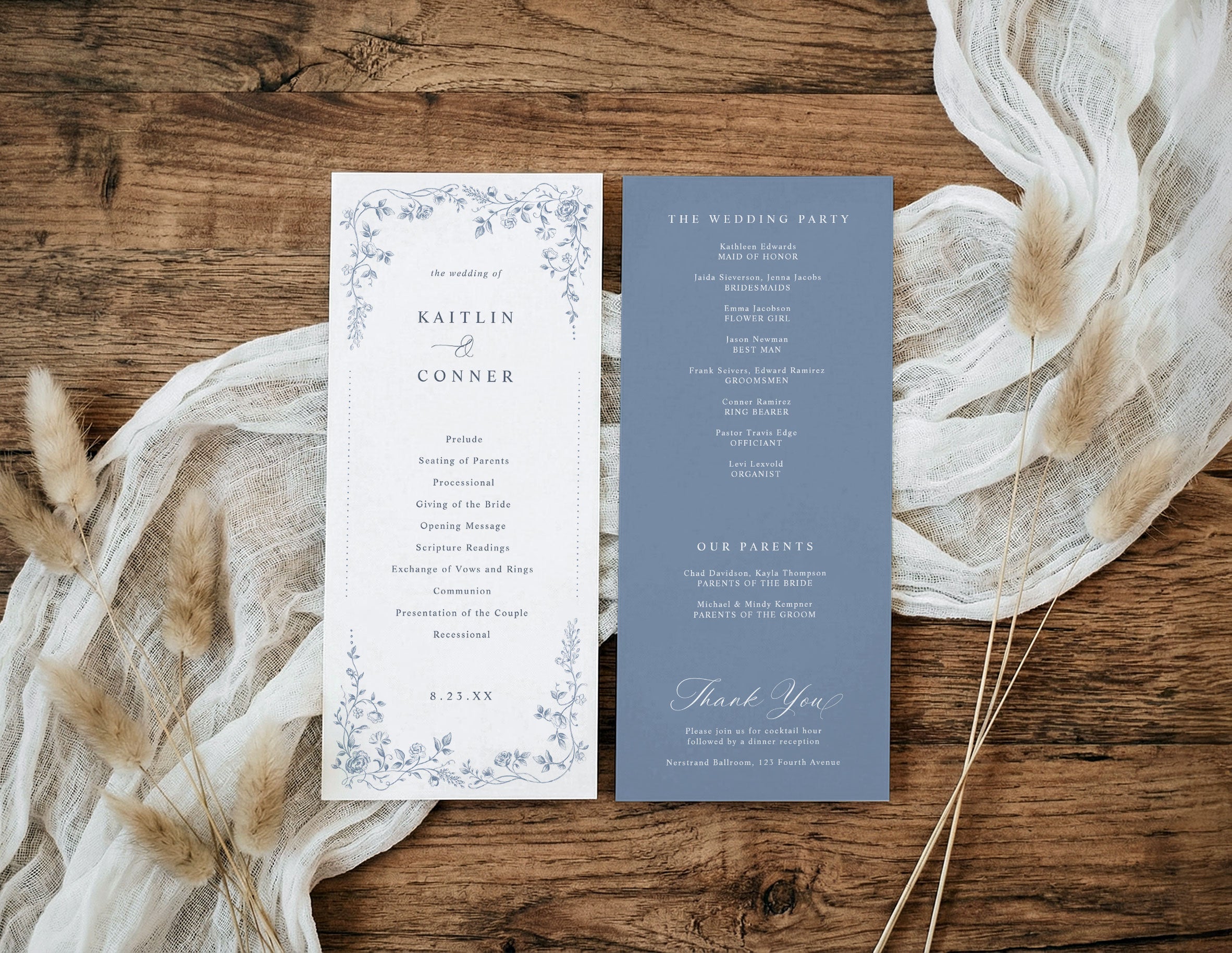 A flat-lay of a wedding program set on a rustic wooden surface, styled with cream gauze fabric and dried pampas grass. The left card is white with a delicate blue floral border and black text, while the right card is a muted slate blue with white text.