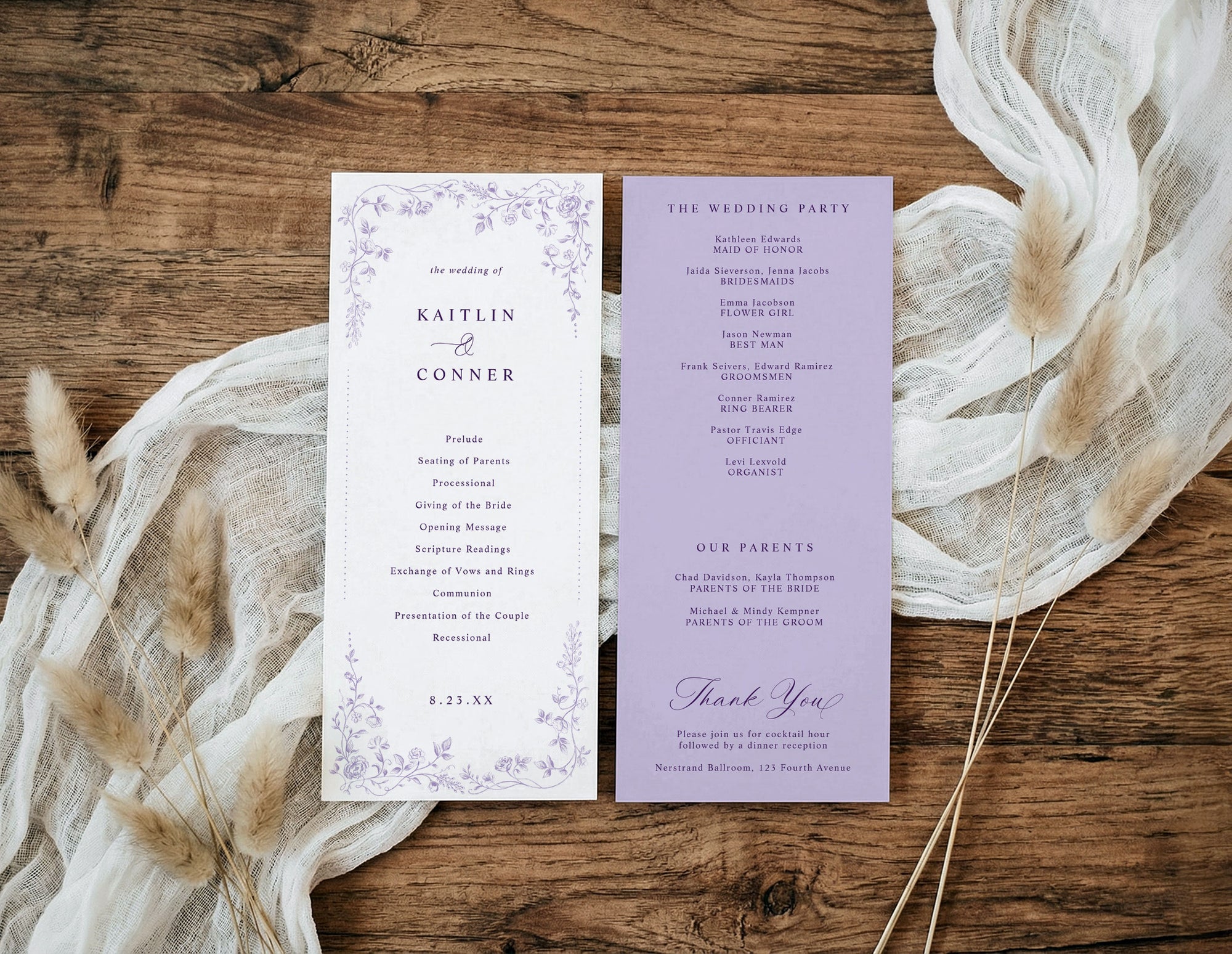 A flat-lay of a double-sided printed wedding program card set on a rustic wooden table, styled with cream gauze and dried pampas grass. The left page is white with a delicate lavender floral border and purple text.