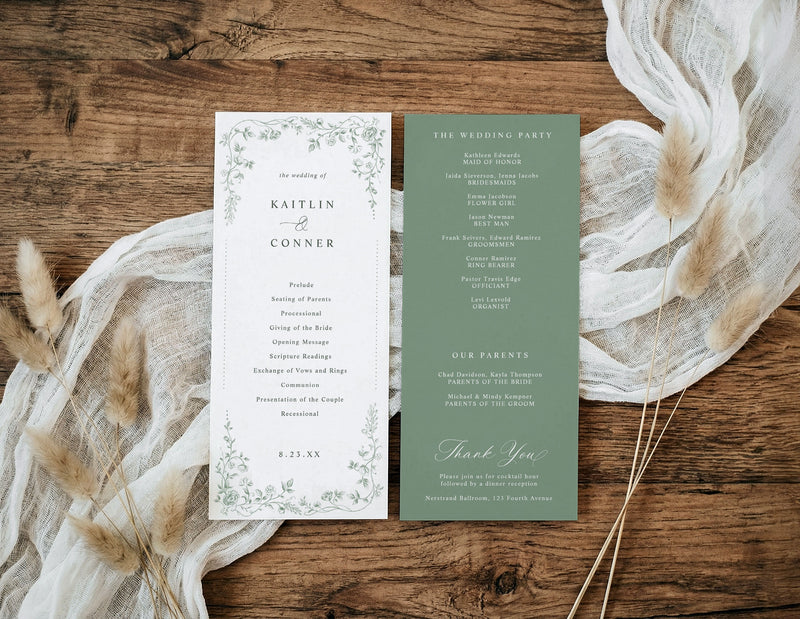 A flat-lay of a double-sided wedding program or invitation set on a rustic wooden surface, styled with cream gauze and dried pampas grass. The left page is white with delicate green floral borders and black text, and the right page is sage green with white text.