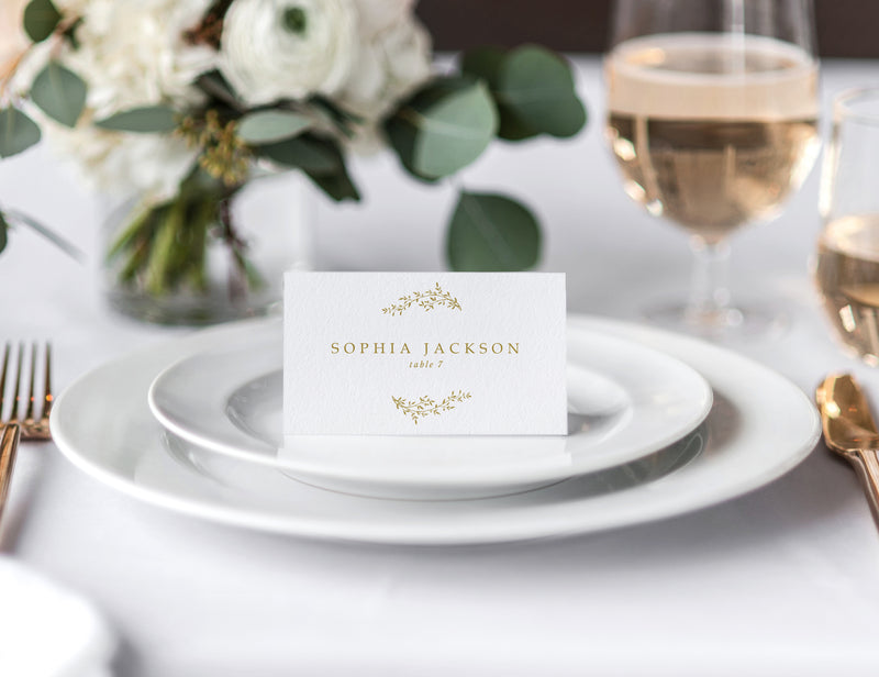 Vintage wedding place card with gold crest and serif font, styled on white dinner plates at elegant table setting