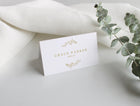 Elegant wedding name card with gold detail and table number, displayed on porcelain plate with floral centerpiece