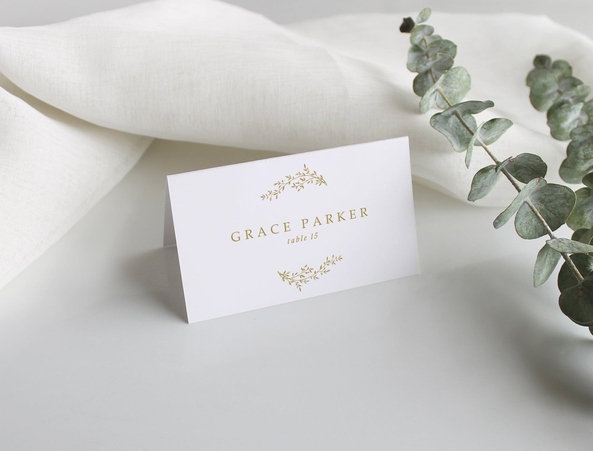 Elegant wedding name card with gold detail and table number, displayed on porcelain plate with floral centerpiece