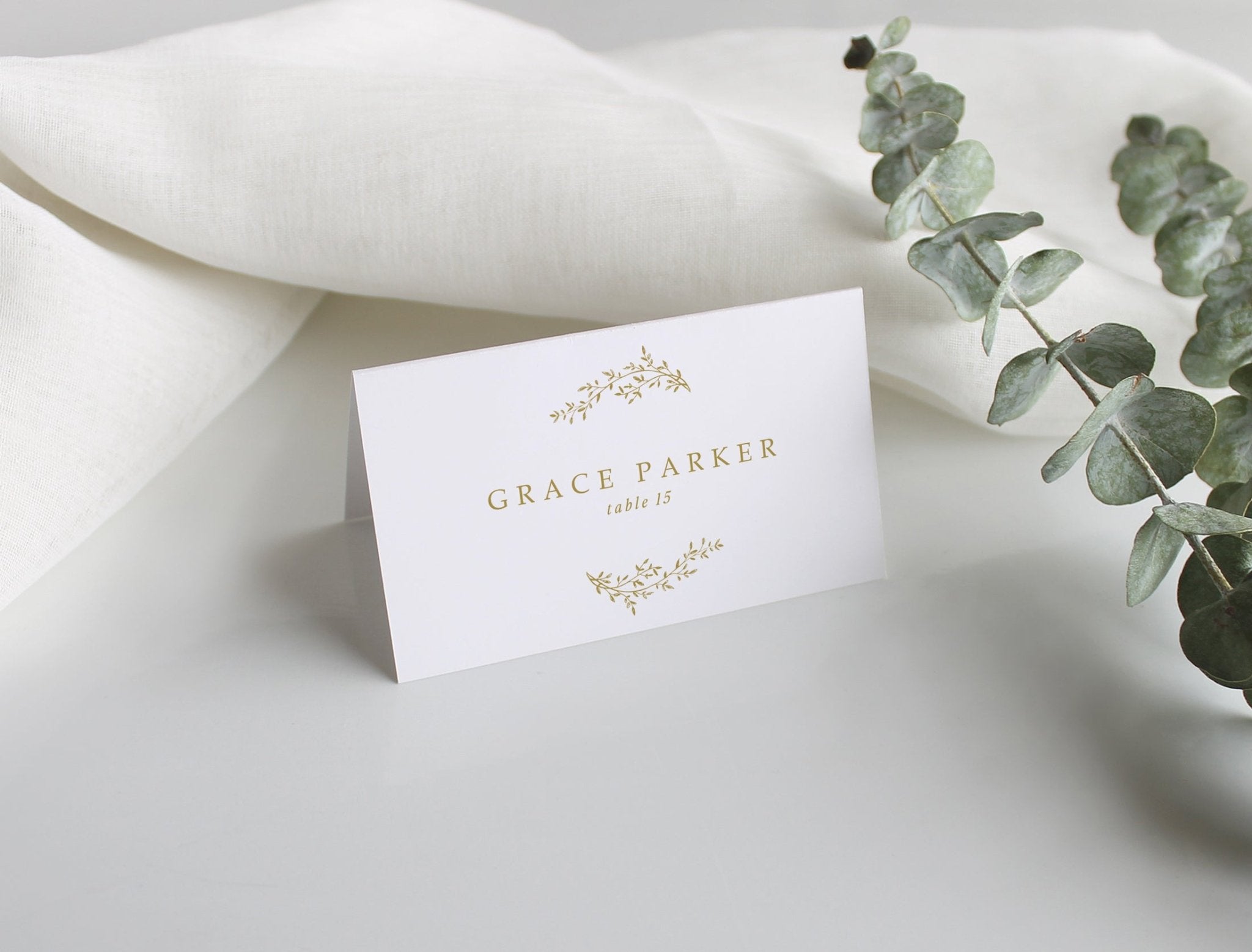 Elegant wedding name card with gold detail and table number, displayed on porcelain plate with floral centerpiece