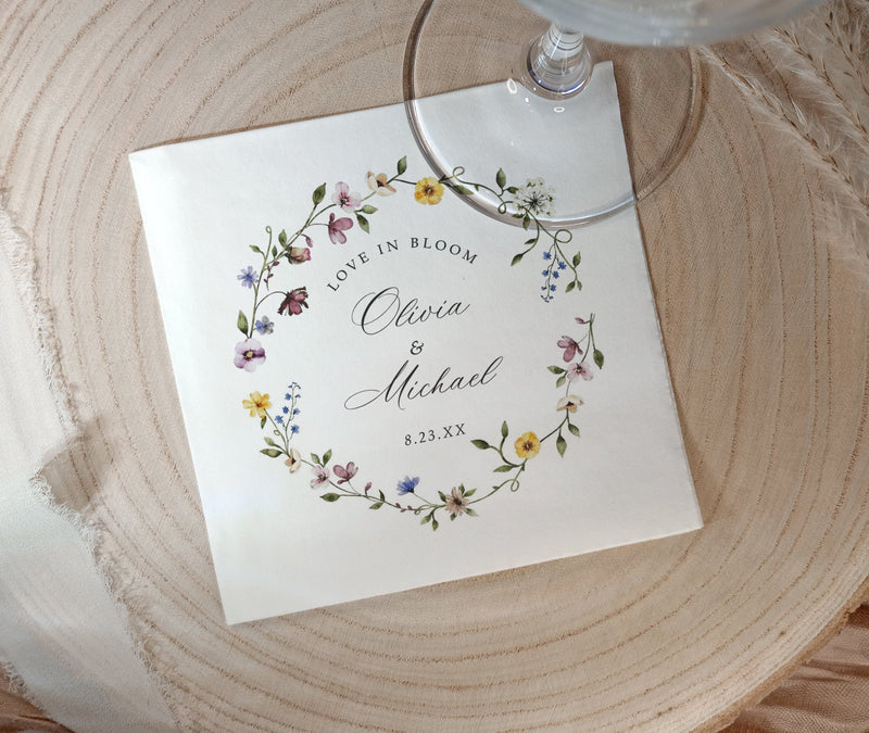 A square white paper bridal shower napkin printed with a floral wreath and the personalized names Olivia & Michael with the date 8.23.