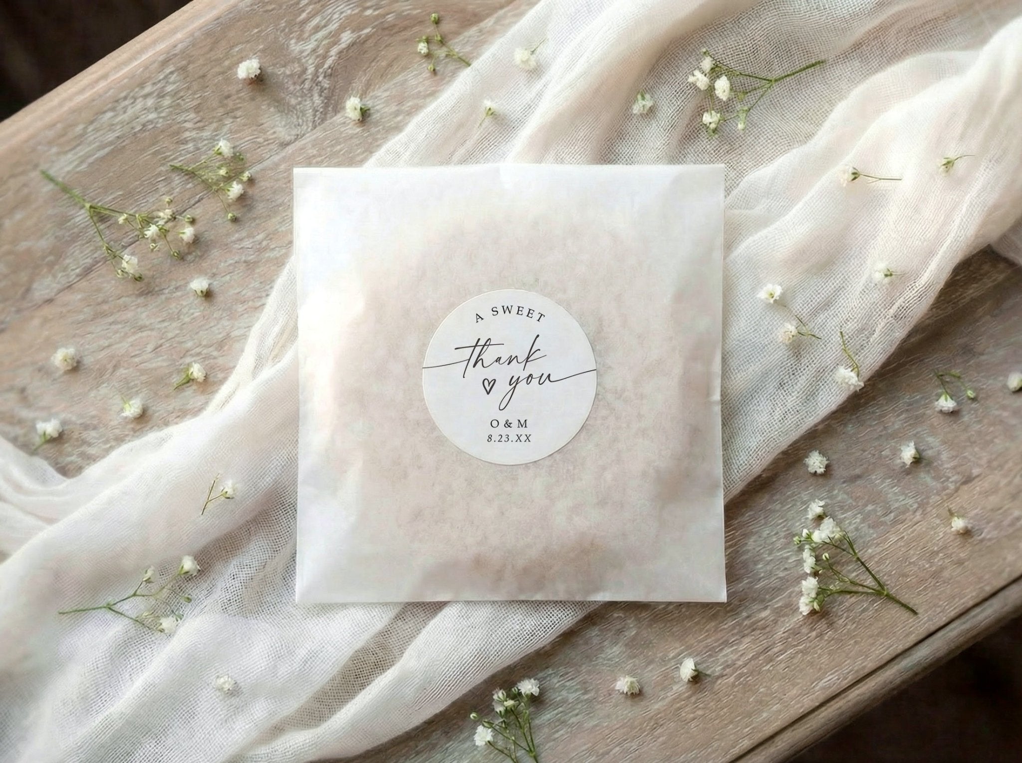 A square white paper wedding favor bag with a round white personalized wedding favor sticker reading 'A Sweet thank you' lies on a rustic wooden surface draped with sheer white fabric and scattered small white flowers.