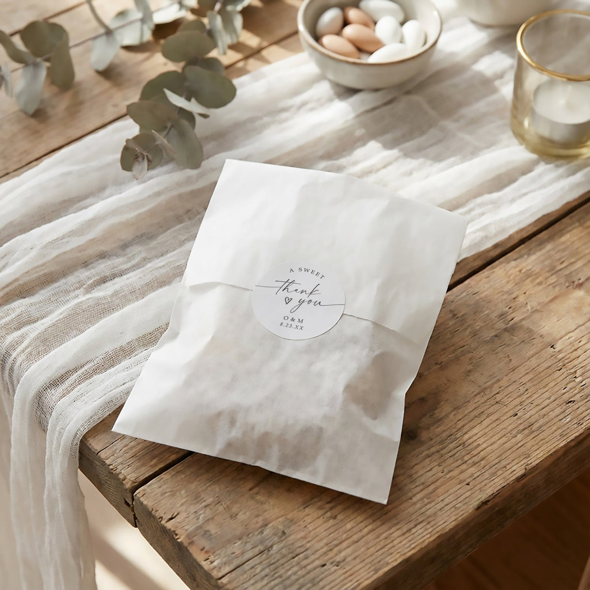 A white paper wedding favor bag with a round printed label reading 'thank you' and 'from your heart' sits on a rustic wooden table draped with sheer white fabric. Decorative eucalyptus stems, a small bowl of candies, and a candle are softly blurred in the background.