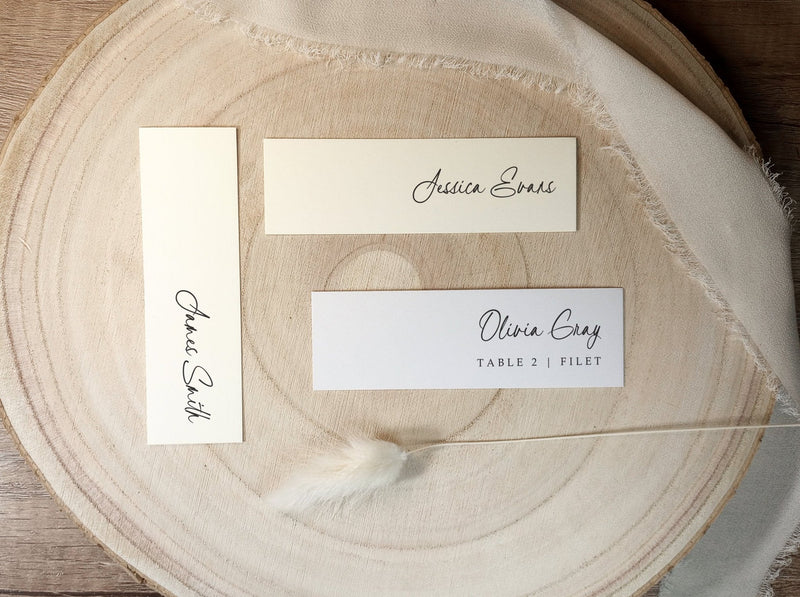 Slim printed wedding name tags with guest names and table numbers, displayed on wood background. Perfect as escort cards or menu accents.
