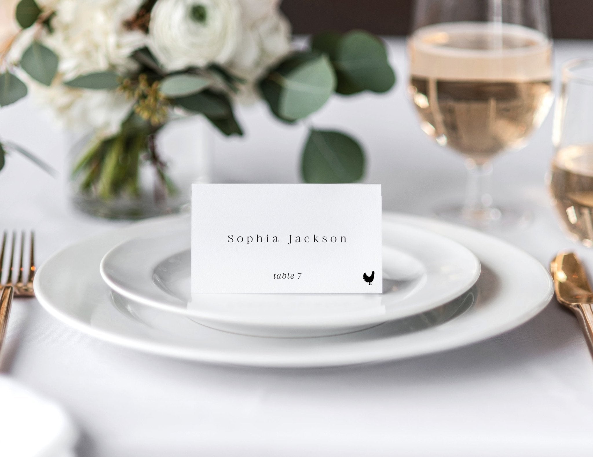 Luxury minimalist wedding place card with guest name and meal icon, displayed on white plate with wine and florals.