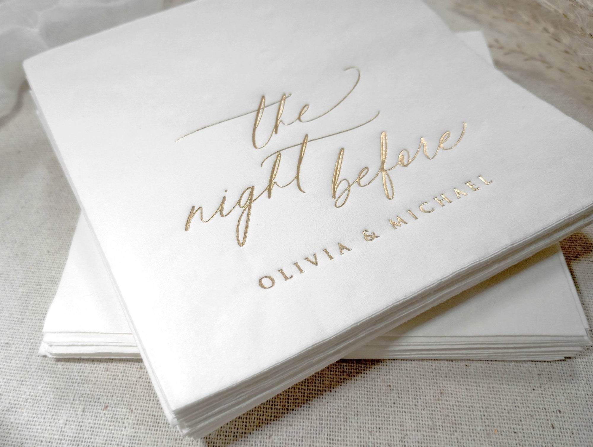 Personalized white cocktail napkin with gold foil names displayed on a linen tablecloth for a wedding rehearsal dinner.