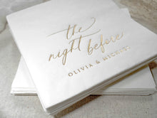 Personalized white cocktail napkin with gold foil names displayed on a linen tablecloth for a wedding rehearsal dinner.