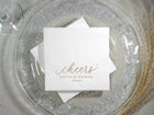 Two custom silver foil “cheers” napkins overlapping on a glass charger plate.