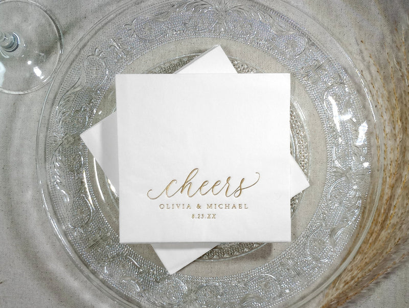 Two custom silver foil “cheers” napkins overlapping on a glass charger plate.