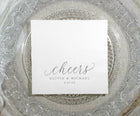 Personalized wedding napkin with “cheers” in silver foil on a clear glass plate