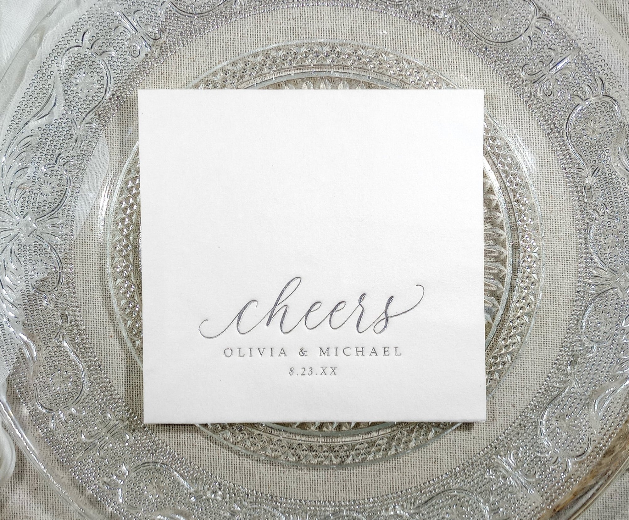 Personalized wedding napkin with “cheers” in silver foil on a clear glass plate
