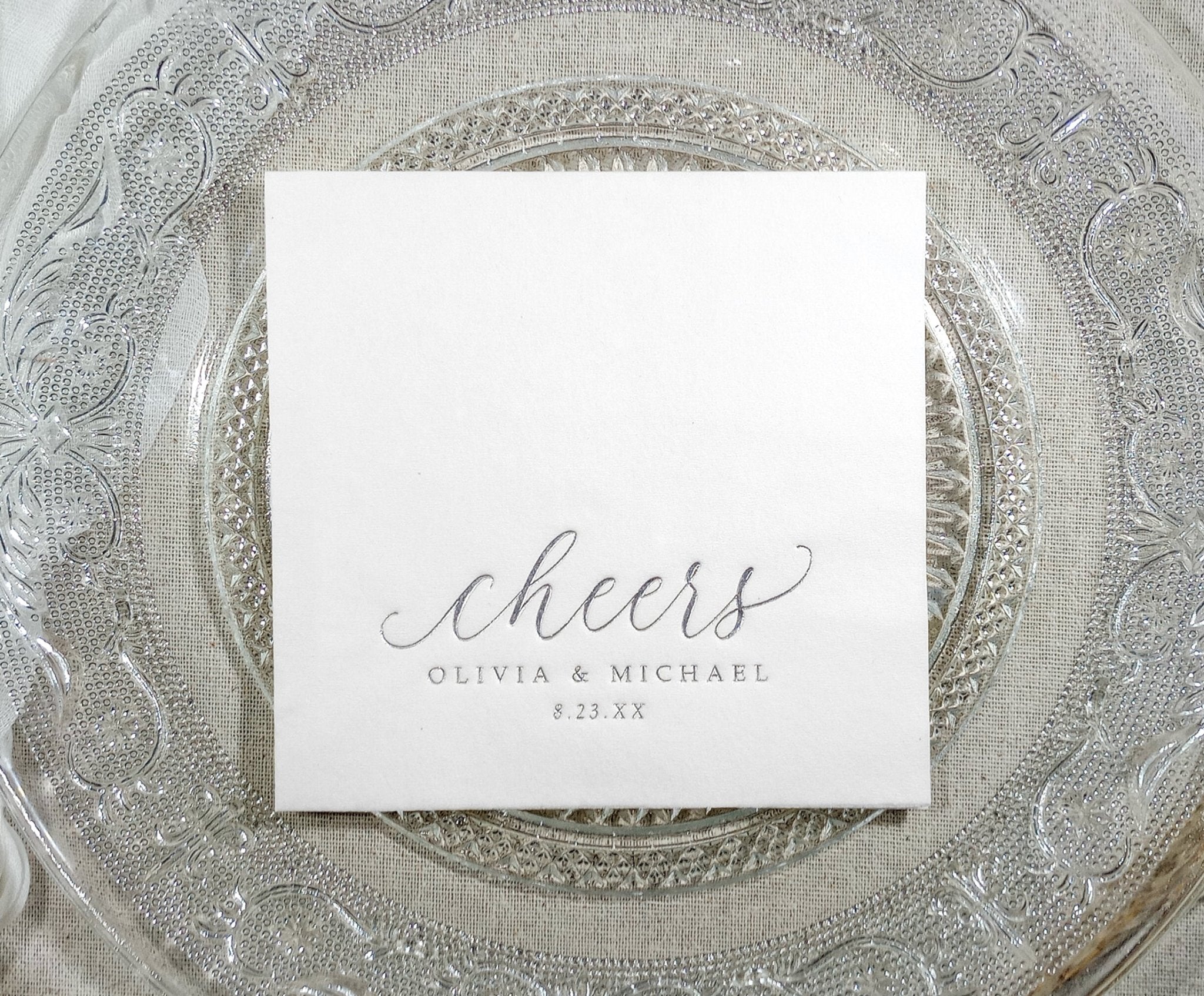 Personalized wedding napkin with “cheers” in silver foil on a clear glass plate