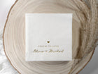 White personalized wedding napkin with gold foil names and wedding date in whimsical script, styled on a blush background.