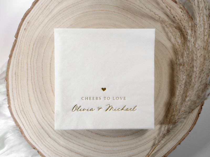 White personalized wedding napkin with gold foil names and wedding date in whimsical script, styled on a blush background.