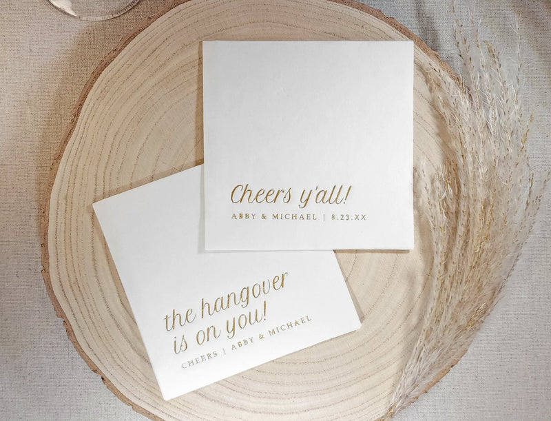 Two gold foil stamped cocktail napkins with gold text on a wooden surface