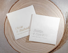Two white foil stamped napkins with gold and silver text on a wooden surface