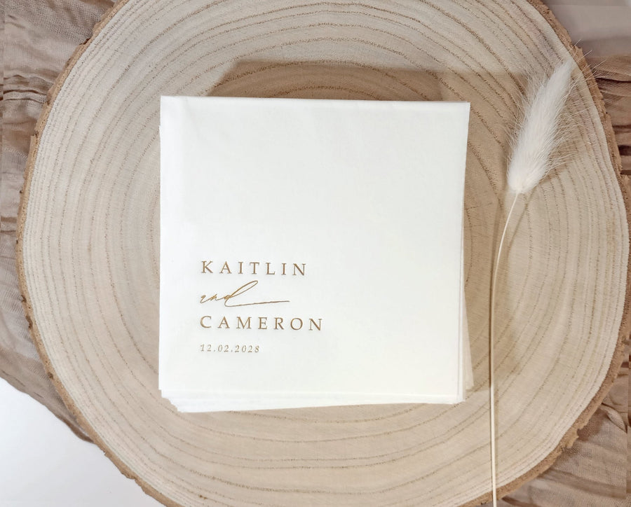 White foil-stamped cocktail napkin with couple's names and wedding date in modern handwritten script.