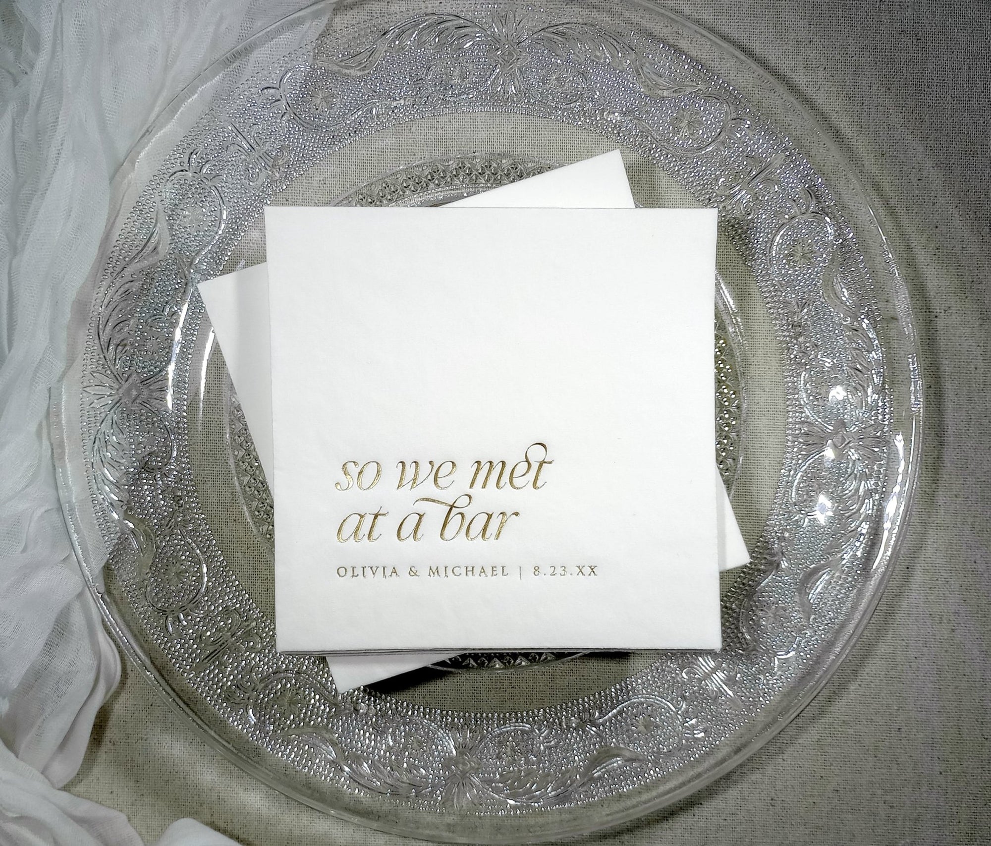 White wedding cocktail napkin with “So We Met at a Bar” in elegant gold foil and custom names below