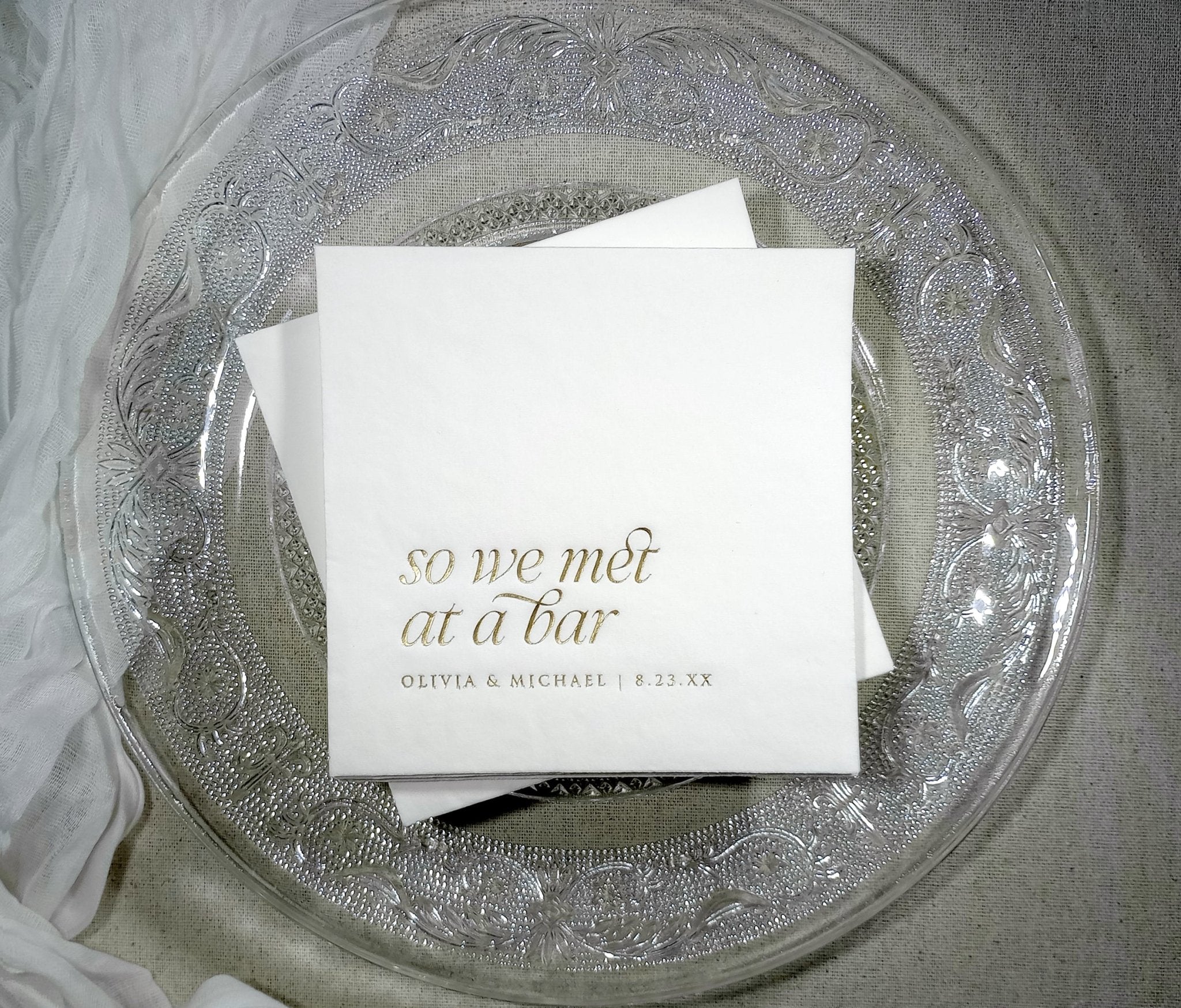 White wedding cocktail napkin with “So We Met at a Bar” in elegant gold foil and custom names below