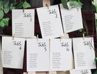Seating arrangement cards for tables with names, pinned to a board with greenery.