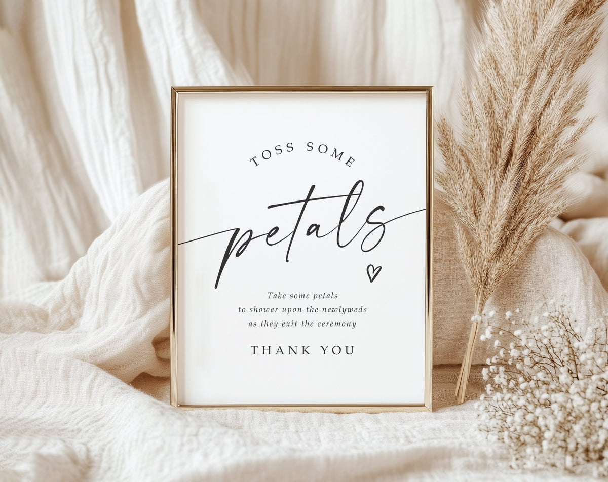 A framed typographic printed wedding sign reading 'Toss some petals' with smaller ceremony text and 'Thank you' below, styled on soft cream fabric with dried pampas grass and baby's breath.