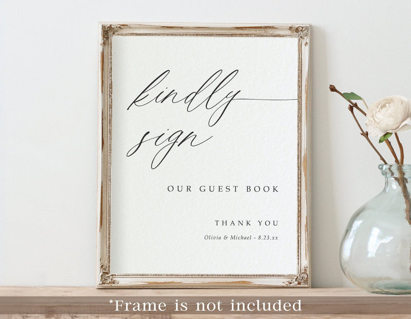 A framed printed guest book sign with elegant black script reading 'kindly sign' and smaller text 'OUR GUEST BOOK' and 'THANK YOU' on a white background. The frame appears distressed and light wood-toned, and the image is styled on a shelf beside a glass vase with a white flower.