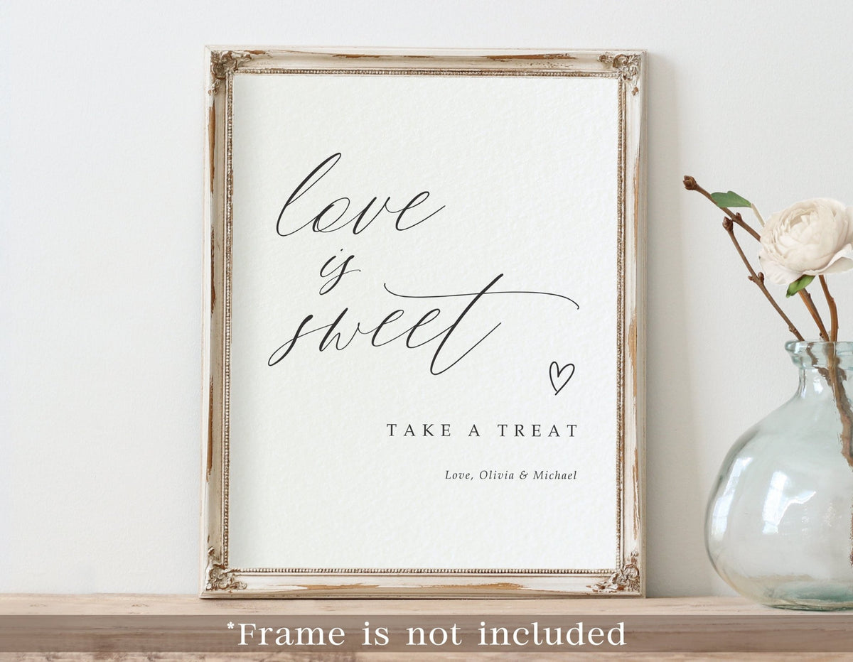 A framed printed wedding sign with the phrase “love is sweet” in black script on a white background, leaning against a light wall on a wooden surface. A small glass vase with a white flower sits to the right.