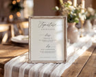 A square framed printed signature cocktails menu sign with elegant black script text on a warm cream background, set on a rustic wooden table with a white striped runner in a softly blurred event venue.