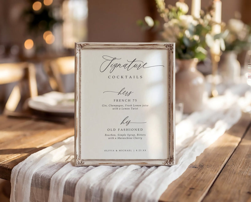 A square framed printed signature cocktails menu sign with elegant black script text on a warm cream background, set on a rustic wooden table with a white striped runner in a softly blurred event venue.
