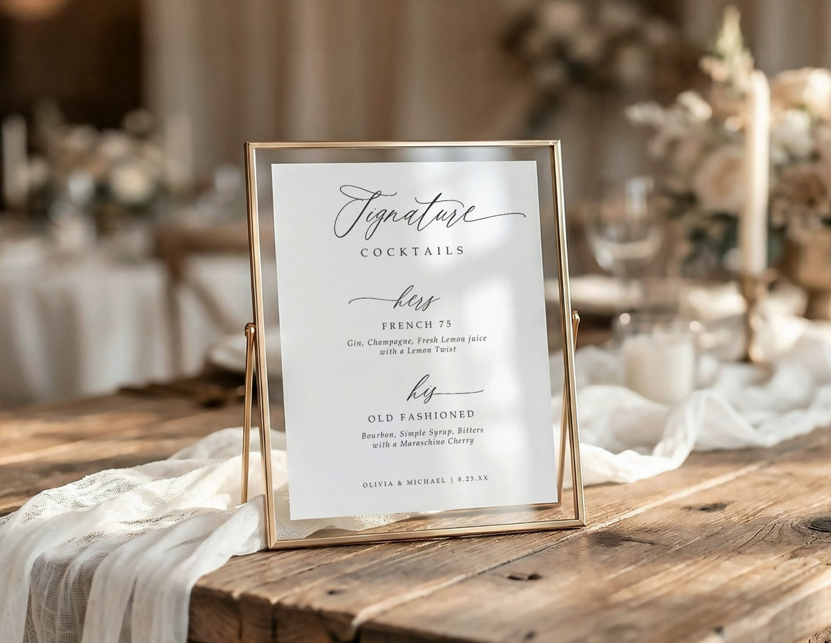 A vertical wedding signature cocktails table sign in a thin gold metal frame, displayed on a rustic wooden table with draped white fabric in a softly blurred event setting.