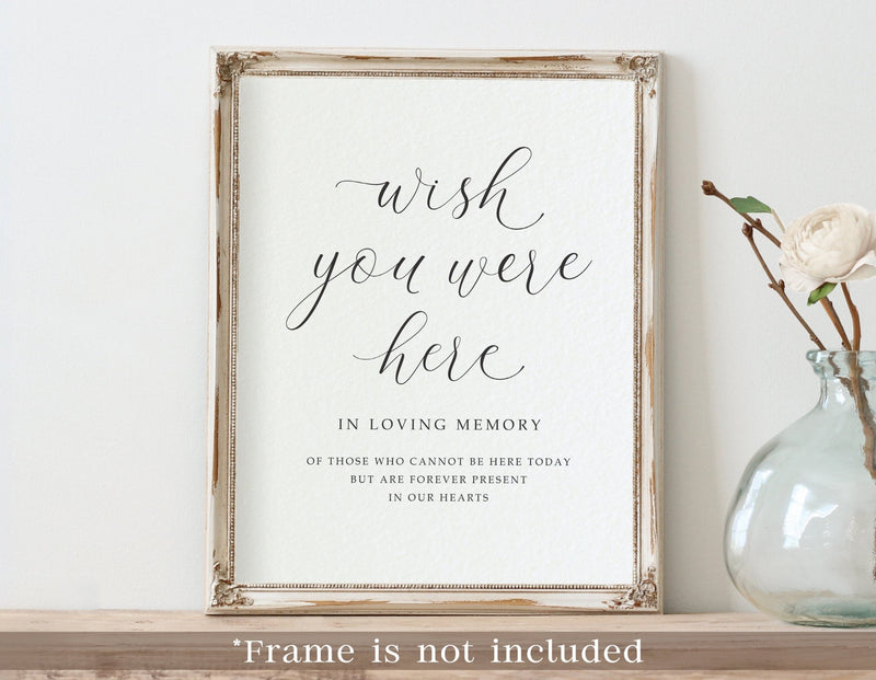A framed custom printed memorial sign with the words 'wish you were here' in black script on a white background, styled on a wooden shelf next to a glass vase with a white flower. The text at the bottom says '*Frame is not included'.